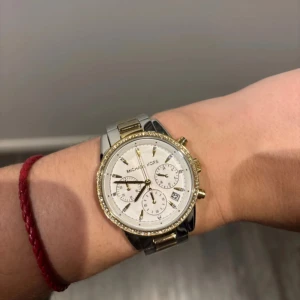 Michael Kors klocka i guld och silver - Snygg Michael Kors klocka med en glittrig boett och vit urtavla. Klockan har en tvåfärgad länkarmband i guld och silver samt flera små urtavlor och datumvisning. Perfekt accessoar för att lyfta din stil.