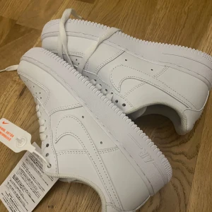 Vita Nike Air Force sneakers - Säljer ett par klassiska vita Nike Air Force sneakers. De har en stilren design med snörning och den ikoniska Nike-loggan på sidan. Perfekta för en casual look.