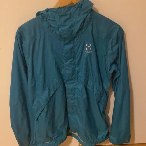 Blå vindjacka från Haglöfs - Säljer en blå vindjacka från Haglöfs med huva och dragkedja. Perfekt för blåsiga dagar och utomhusaktiviteter. Jackan har långa ärmar och en enkel design.