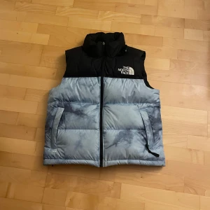 Dunväst från The North Face - Snygg väst från The North Face i svart och ljusblått splitter ny köpt för mamman men hon hade redan en sådan Och tänkte därför sälja denna. Perfekt för kyligare dagar och har en stilren design med logotyp på bröstet. Passar bra på 165-170cm långa!!