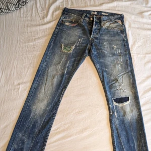 Blå jeans från Replay - Säljer ett par blå Replay jeans med slitna detaljer och hål för en trendig look. Perfekta för en avslappnad stil.