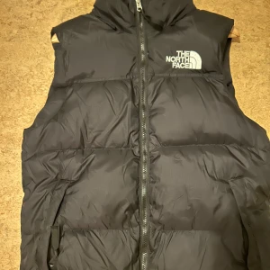Svart dunväst från The North Face - Snygg och praktisk svart dunväst från The North Face med dragkedja framtill. Perfekt för kyligare dagar och har en klassisk design med logotyp på bröstet och ryggen.