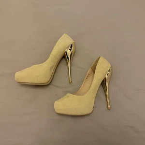 Beige pumps med gulddetaljer - Eleganta beige pumps med hög klack i guld. Perfekta för att ge en lyxig touch till din outfit. Skorna har en rundad tå.