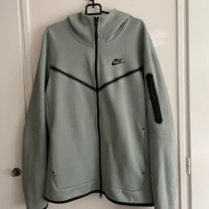 Grön huvtröja från Nike - Säljer en stilren grön huvtröja från Nike med dragkedja och huva. Tröjan har en ficka med dragkedja på ärmen och två sidofickor. Perfekt för en sportig look.