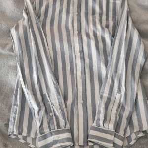 Randig skjorta från Pier One - Helt oanvänd snygg randig skjorta från Pier One i blått och vitt. Skjortan har långa ärmar och knappar framtill samt en bröstficka. Perfekt för en stilren look.