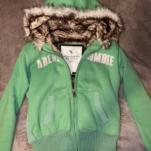 Grön hoodie från Abercrombie & Fitch - Säljer en grön hoodie från Abercrombie & Fitch med mjukt foder i luvan och dragkedja framtill. Den har ribbade muddar och en snygg logga på bröstet. Perfekt för kyliga dagar!