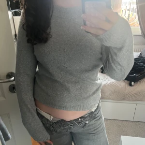 Grå stickad tröja - Säljer en mysig grå stickad tröja med långa ärmar. Perfekt för kyligare dagar och enkel att matcha med olika outfits. Tröjan har en klassisk design med ribbstickade muddar vid ärmslut och nederkant.