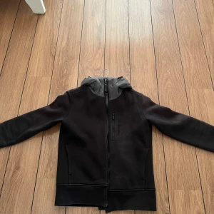 Svart jacka med huva - Snygg svart jacka med grå huva och dragkedja framtill. Den har en stilren design med långa ärmar och en bekväm passform. Säljs pga har blivit för liten, den är i barn storlek m och är lite sol blekt men inget man tänker på