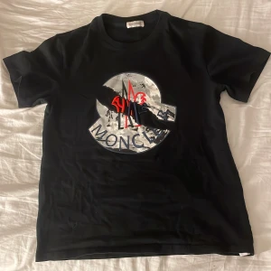 Svart t-shirt från Moncler - Snygg svart t-shirt från Moncler med ett stort, iögonfallande tryck på framsidan. Perfekt för en stilren och modern look. Passar bra till både jeans och shorts.