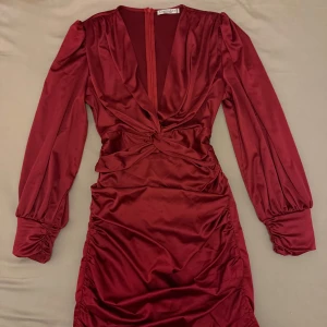 Vinröd klänning med knytdetalj - Elegant vinröd klänning i satin med långa ärmar och en v-ringad design. Klänningen har en knytdetalj framtill och en dragkedja i ryggen. Perfekt för festliga tillfällen.
