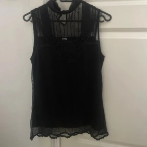 Svart ärmlös blus med spetsdetaljer - Elegant svart ärmlös blus med spetsdetaljer och hög hals. Perfekt för en stilren look med transparenta partier och dekorativ spets vid halsen. Från bikbok i storlek s