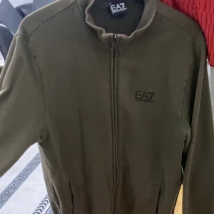 Olivgrön jacka från EA7 - Snygg olivgrön jacka från EA7 med dragkedja framtill. Jackan har en stilren design med långa ärmar och två fickor. Perfekt för en casual look.