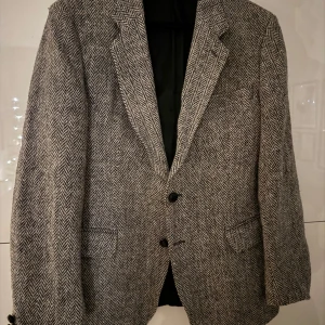 Grå Harris Tweed kavaj från Grosvenor House - Säljer en stilren grå kavaj från Grosvenor House, tillverkad av 100% ull från Harris Tweed. Kavajen har ett klassiskt fiskbensmönster och två knappar framtill. Perfekt för en elegant look.
