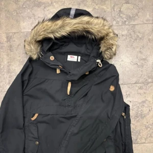 Fjällräven jacka - Fjällräven Iceland anorak dam. Svart parkajacka från Fjällräven med pälsdetaljer på huvan. Jackan har flera praktiska fickor och knappar samt en dragkedja framtill. Perfekt för kyliga dagar. Använd fåtal gånger. (Har inga fler bilder för tillfället)