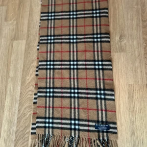 Rutig halsduk från Burberry - Snygg och klassisk rutig halsduk från Burberry i beige, svart, vit och röd. Tillverkad i mjukt material med fransar i ändarna. Perfekt för att hålla dig varm och stilren under kyliga dagar.