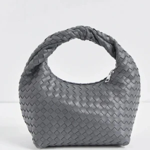 Grå flätad handväska - Snygg grå handväska med flätat mönster och dragkedja. Perfekt för att ge din outfit en stilren touch. Väskan har ett unikt handtag som också är flätat, vilket ger en extra detalj till designen.
