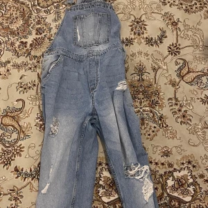 Blå jeansbyxdress med slitningar - Snygg blå jeansbyxdress med slitningar för en trendig look. Byxdressen har en klassisk design med hängslen och en ficka på bröstet. Perfekt för en avslappnad stil.