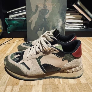 Valentino Rockrunner sneakers i camouflage - Snygga Valentino Rockrunner sneakers med ett coolt camouflagemönster i grönt och beige. Skorna har en unik design med röda detaljer på hälen och snörning framtill. Skicket 7,5/10 finns hål i hälen stl 39/5 passar även 40 annars i bra skick box ingår såklart med lite snören skriv för fler frågor mvh 😊