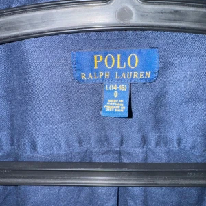Mörkblå skjorta från Ralph Lauren - Snygg mörkblå skjorta från Ralph Lauren med klassisk krage och knappar framtill. Den har en broderad röd logotyp på bröstet och långa ärmar. Perfekt för en stilren look. Ny pris 1199kr. Passar 14/16 år. Använd endast 1 gång