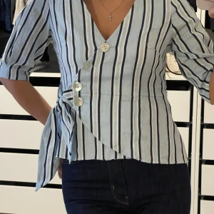 Randig omlottblus med knyt Zara - Säljer en stilren randig omlottblus från H&M i ljusblått och vitt. Blusen har trekvartsärmar och en v-ringad design med knappar framtill och ett knytband i midjan. Perfekt för en chic och avslappnad look.