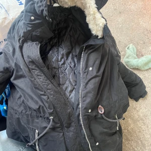 Svart parkajacka med fuskpäls - Säljer en svart parkajacka från Divided med fuskpälsfoder i huvan. Jackan har dragkedja och knappar framtill samt justerbar midja med snörning. Perfekt för kyliga dagar!