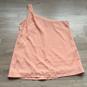 Persikofärgad oneshoulder topp från Nelly - Säljer en stilren persikofärgad oneshoulder topp från Nelly. Toppen har en enkel design med en axelrem och är perfekt för varma dagar. Materialet har en lätt skimrande design. Passar utmärkt till både jeans och kjol. En liten tråd som gått upp nertill, se sista bilden, inget som syns på håll eller lägga märke till. 
