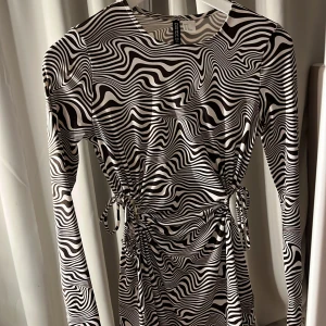 Svartvit mönstrad klänning  - Säljer en cool svartvit klänning  med ett unikt vågmönster. Klänningen har långa ärmar och snörning i midjan för en justerbar passform. Perfekt för att sticka ut med stil!