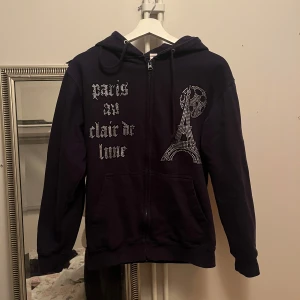 Mörkblå hoodie med Paris-motiv från Blinglucid - Säljer en snygg mörkblå hoodie från Blinglucid med dragkedja och ett glittrigt Paris-motiv på framsidan. Hoodien har långa ärmar och en justerbar huva med snören.