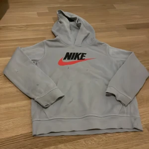 Grå hoodie från Nike - Säljer en grå hoodie från Nike med det klassiska logotyptrycket i svart och rött på framsidan. Tröjan har en bekväm huva och långa ärmar. Perfekt för en avslappnad stil.