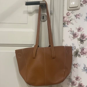 Brun axelväska  - Snygg brun axelväska i skinn med stilrena sömmar och både långa och handtag. Perfekt för att bära med sig det nödvändigaste. Väskan har en rymlig insida och en enkel design som passar till många tillfällen. Liknar väskan från Gina. Har inte använt den alls!