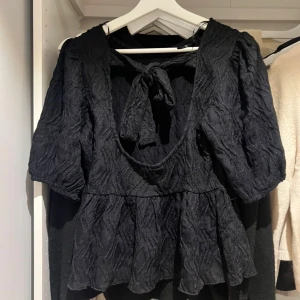 Svart topp med knyt och volang - Snygg svart topp med knyt i ryggen och volangdetaljer. Toppen är från Gina i storlek Xs