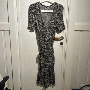 Svartvit mönstrad klänning från indiska  - Snygg svartvit klänning från indiska med ett abstrakt mönster. Klänningen har en v-ringad design och korta ärmar. Den är knytbar i midjan för en smickrande passform. Perfekt för både vardag och speciella tillfällen.