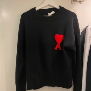 Svart tröja från Ami Paris - Säljer en stilren svart tröja från Ami med ett rött hjärtmotiv på bröstet. Tröjan har långa ärmar och en rund halsringning. Perfekt för en trendig look.