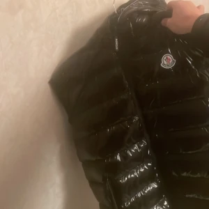 Svart puff väst från Moncler  - Säljer en svart pufferväst  från Moncler med glansig yta och ikonisk logga på bröstet. Jackan har en dragkedja framtill och är perfekt för kyligare väder. Insidan har ett unikt serietidningsmönster som ger en cool touch. Storlek l men pass både m L
