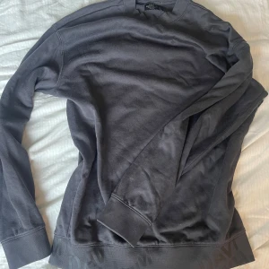 Mörkgrå sweatshirt från H&M (David beckham kollektion) - Säljer en stilren mörkgrå sweatshirt från David Beckham. Tröjan har långa ärmar och en rund halsringning. Perfekt för en avslappnad stil. Textdetaljer vid nederkanten ger en extra touch.