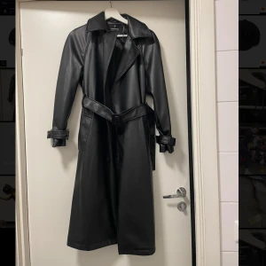 Svart trenchcoat från Stradivarius - Snygg svart trenchcoat i syntetmaterial från Stradivarius.. Jackan har ett bälte i midjan och långa ärmar med detaljerade manschetter. Perfekt för en stilren look. Endast kommit till användning 1 gång.
