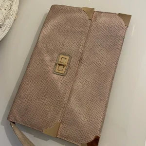 Beige kuvertväska med gulddetaljer - Elegant beige kuvertväska med guldfärgade hörndetaljer och ett stilrent lås framtill. Väskan har en diskret ormskinnsmönstrad yta och en praktisk handledsrem. Insidan är rymlig med fickor för småsaker. Perfekt för en chic look!  Längd:22,22cm och bred : 33cm