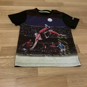 Svart t-shirt med fotbollstryck - Cool svart t-shirt med ett stort fotbollstryck på framsidan som visar en actionfylld matchscen. Perfekt för fotbollsfans! T-shirten har korta ärmar och en rund halsringning.