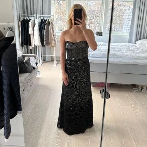 Svart glittrig långklänning - Säljer en elegant svart långklänning med glittrande detaljer. Klänningen är axelbandslös och har en figurnära passform som ger en stilren siluett. Perfekt för en festlig kväll eller bal. Märket är Panos Emporio. Storlek 38, M. Nypris är 2499kr. Den har dold dragkedja i sidan