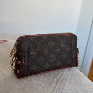 Brun monogram handväska/axelremsväska med kedja - Snygg brun multifunktionell väska med klassiskt monogrammönster och guldfärgad kedja. Väskan har en dragkedja upptill och detaljer i rött längs kanterna. Tillhörande extra handtag tillkommer!