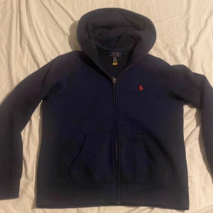 Mörkblå hoodie från Ralph Lauren - Säljer en snygg mörkblå hoodie från Ralph Lauren med dragkedja och det klassiska röda broderade logotypen på bröstet. Perfekt för en avslappnad stil. Den har långa ärmar och en praktisk huva.