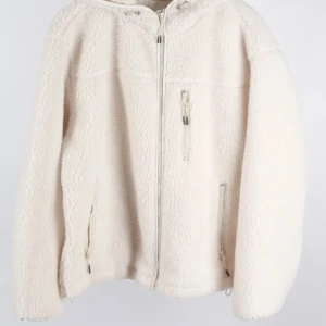 Vit teddyjacka med dragkedja H&M - Mysig vit teddyjacka med dragkedja framtill och praktiska fickor. Perfekt för kyliga dagar med sin fluffiga och varma design. Jackan har långa ärmar och en hög krage för extra värme.