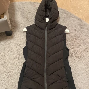 Svart dunväst från Zara - Snygg svart dunväst från Zara med huva och dragkedja. Perfekt för kyligare dagar med sin quiltade design som ger extra värme och stil. Västen har en normal passform och är ett mångsidigt tillskott till garderoben, använt ett par gånger och storleken är S/XS! Kan tänka mig gå ner till 200/250kr