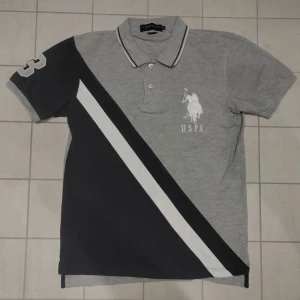 Grå och svart pikétröja från U.S. Polo Assn. - Snygg pikétröja från U.S. Polo Assn. i grått och svart med vit diagonal rand. Tröjan har korta ärmar och en klassisk krage med knappar. Märkets logga är broderad på bröstet. Perfekt för en stilren look.