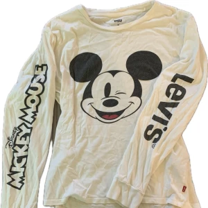 Vit långärmad tröja med Disney tryck från Levi's - Säljer en vit långärmad tröja från Levi's med en stor Mickey Mouse-tryck på framsidan. Limited edition tröja som inte sälj mer, super skön, en toppen mys tröja. Tröjan har texten 'Mickey Mouse' och 'Levi's' tryckt på ärmarna. Perfekt för Disney-fans!