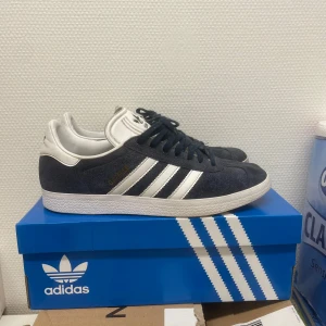 Adidas Gazelle i mörkblå mocka - Snygga Adidas Gazelle sneakers i mörkblå mocka med vita detaljer och snörning. Klassisk design med de ikoniska tre ränderna på sidan. Perfekta för en stilren och avslappnad look. Box ingår.