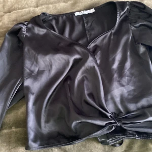 Svart satinblus från NA-KD - Elegant svart blus i satin från NA-KD med knytning framtill. Blusen har långa ärmar och en v-ringad design som ger en stilren look. Perfekt för en chic och sofistikerad stil. 