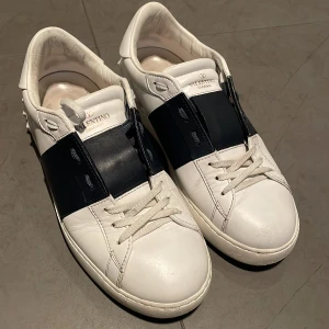 Valentino Garavani sneakers i vitt och blått - Snygga Valentino Garavani sneakers i vitt skinn med en bred blå rem över snörningen. Skorna har en stilren design med dekorativa nitar på hälen. Perfekta för en trendig look. Har inte använts och sulan är fortfarande jätte bra!💗storlek 39.5