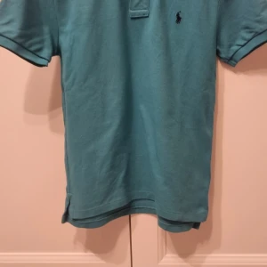 Turkos pikétröja från Ralph Lauren - Snygg turkos pikétröja från Ralph Lauren med klassisk krage och korta ärmar. Tröjan har en liten broderad logga på bröstet. Perfekt för en avslappnad och stilren look.