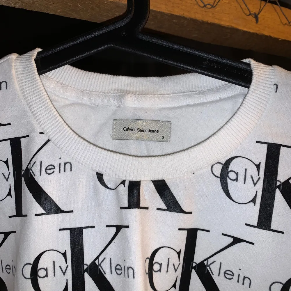 Säljer en vit tröja från Calvin Klein med ett stort, svart logotypmönster över hela plagget. Tröjan har långa ärmar och en rund halsringning. Perfekt för dig som gillar en stilren men ändå iögonfallande look. Passar bra till jeans eller kjol för en avslappnad stil.. Neuletakit & Villapaidat.